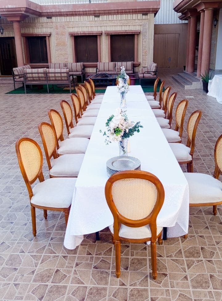 طاولة خارجية طويلة بمفرش أبيض وكراسي كين